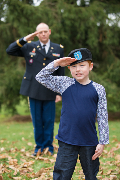 Army son wearing Dad's beret and saluting the flag with Dad blurry in background saluting the flag