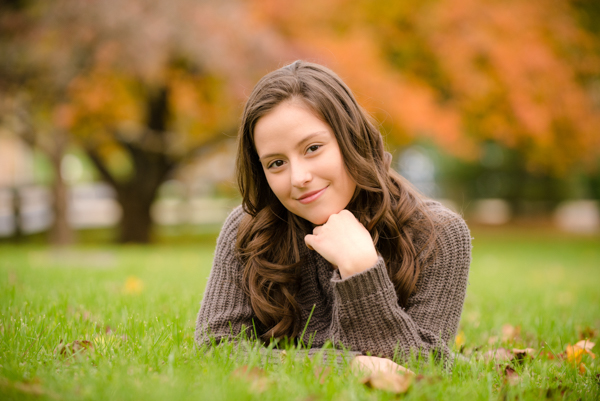 high school senior girl laying in grass with fall leaves blurry in the background
