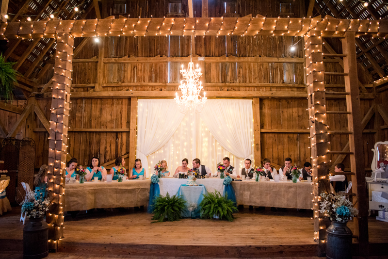 Lit up platform inside an old rustic barn with chandelier hanging above the head table and lights wrapped around banisters. Taken by J & D Studio, Harrisburg Photographers